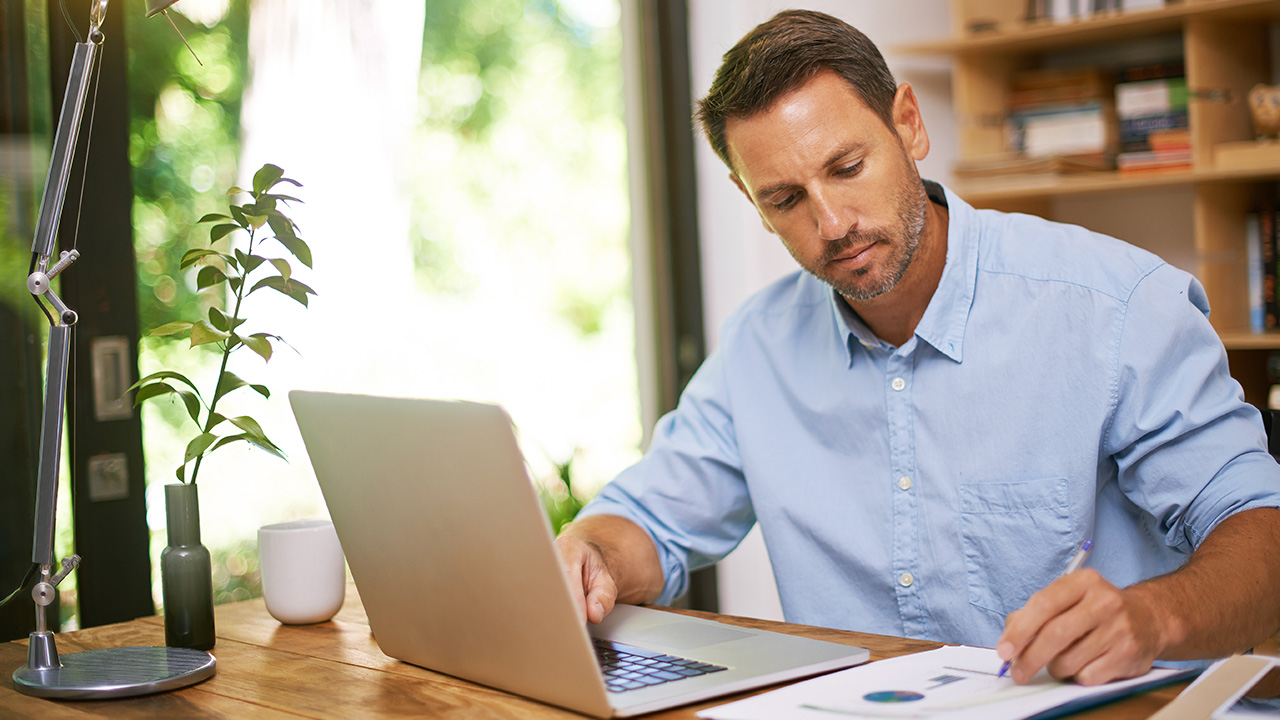 A writing down on his paper while working on his laptop
